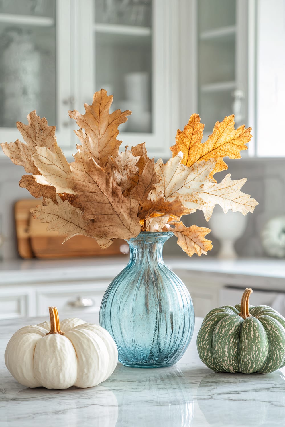 An elegant autumnal still life with a blue vase containing brown and gold fall leaves, flanked by a white pumpkin on the left and a green pumpkin on the right, set on a polished marble countertop against a backdrop of white cabinetry and a blurred kitchen scene.