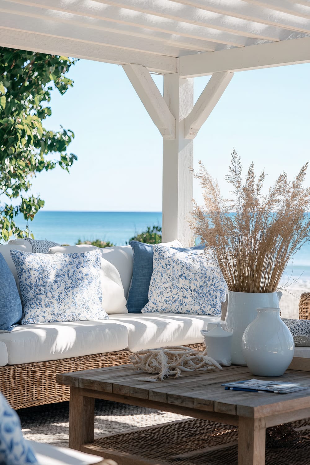 An outdoor seating area under a white pergola, overlooking a serene beach and blue sea. The setup includes a wicker sofa adorned with white and blue-patterned cushions, and a wooden coffee table decorated with white ceramic vases and a bundle of dried ornamental grass. The scene is bright and airy, with natural sunlight casting gentle shadows.