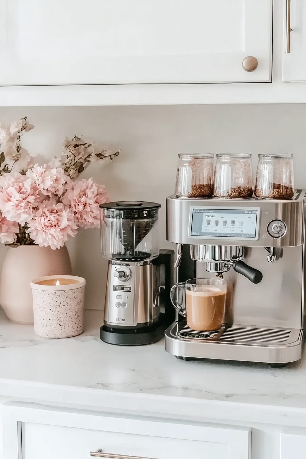 A modern kitchen countertop featuring a stainless steel espresso machine with a digital screen, a ceramic cup filled with coffee, a coffee grinder, a candle in a speckled container, and a vase with pink flowers.