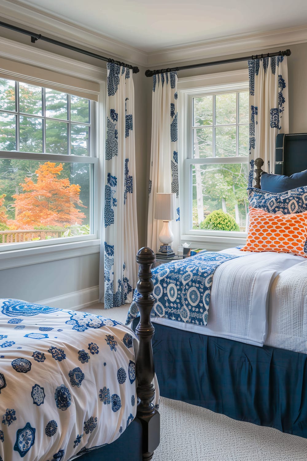 A well-lit bedroom featuring a pair of twin beds adorned with blue and white patterned bedding. Each bed has a dark wooden post frame. The room has large windows with white curtains, accented with blue floral patterns, allowing natural light to flood in. A white table lamp stands between the beds, placed on a small nightstand. Outside the windows, lush green trees and a tree with vibrant orange foliage create a picturesque view.