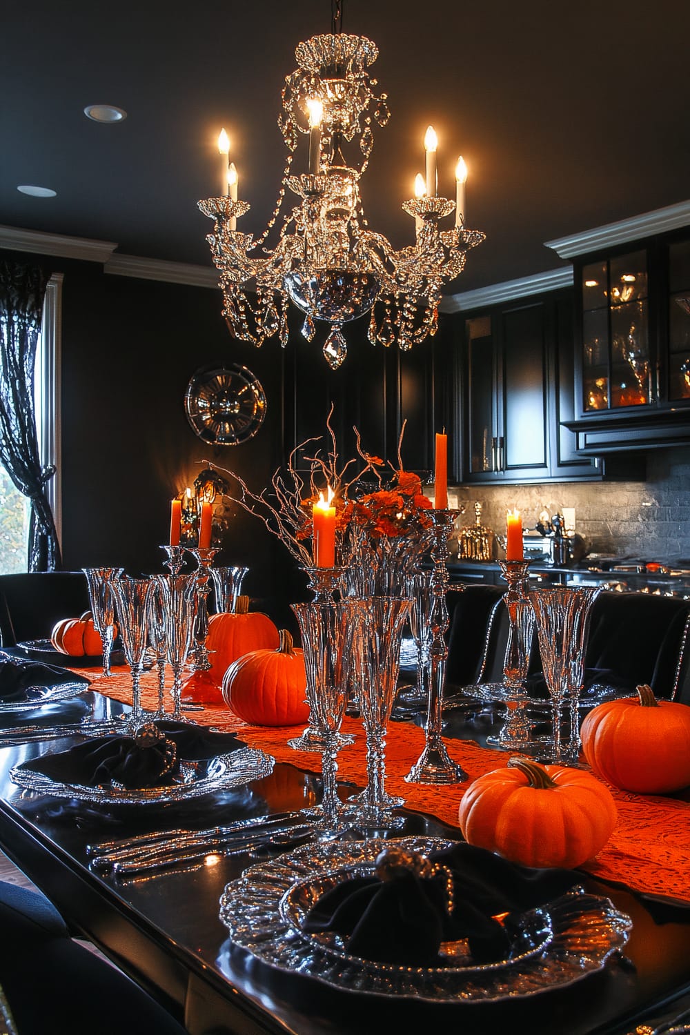 A lavishly set dining table is adorned with sparkling crystal glassware, elegant candle holders with orange candles, and small pumpkins. The table runner is a bold orange, adding a vibrant contrast to the dark, sophisticated setting. Overhead, a grand chandelier with ornate crystals illuminates the scene, reflecting light off the glass and metal surfaces. The background features dark cabinetry and modern kitchen fixtures, enhancing the moody and opulent atmosphere.