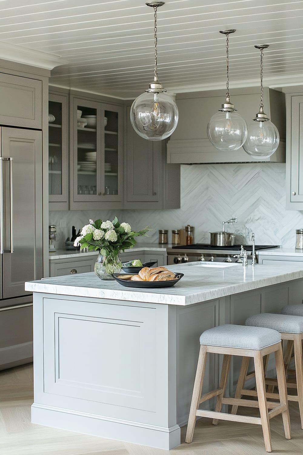 A sophisticated kitchen interior featuring a central island with a white marble countertop. On the island are a vase of fresh flowers and a platter with pastries. Above the island, three spherical glass pendant lights with metal fixtures hang from the ceiling. The cabinetry is painted in a soft grey color, with upper cabinets featuring glass-front doors that reveal neatly stacked dishes inside. The backsplash is a chevron-patterned marble tile, complementing the stainless-steel appliances including a refrigerator and a stove. The island is paired with three counter stools with light grey upholstered seats and wooden legs.