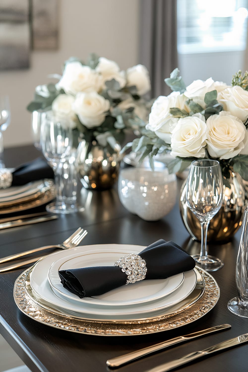 Elegant dining table setting featuring white plates with gold-rimmed borders, accompanied by black napkins encircled by sparkling jeweled rings. The table is subtly adorned with white rose arrangements in gold vases, clear wine glasses, and silverware arranged in a polished manner. The background includes blurred wall art and draped curtains, highlighting the sophisticated ambiance of the setting.