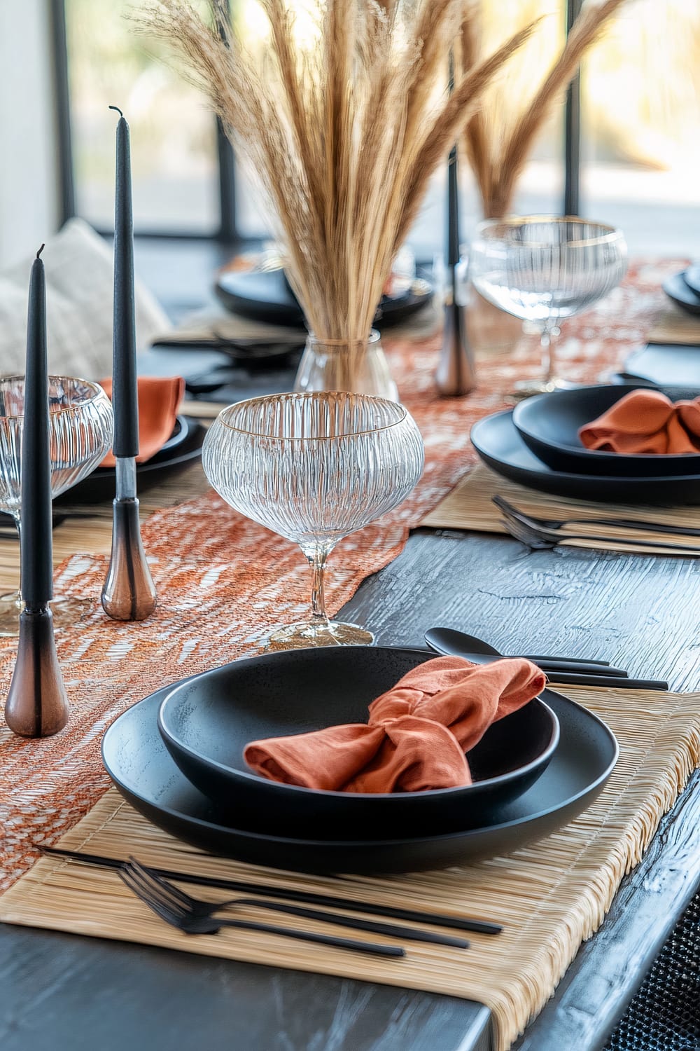 A sophisticated and modern dining table setting is beautifully adorned with black ceramic plates and bowls, each complemented by burnt orange cloth napkins. Glass goblets with a ribbed texture enhance the elegance, while tall, slender black candles in metallic holders bring a dramatic and intimate ambiance. Natural beige placemats add texture, and the table runner in rich orange tones, combined with a centerpiece of dried pampas grass, completes the autumnal, minimalist aesthetic.