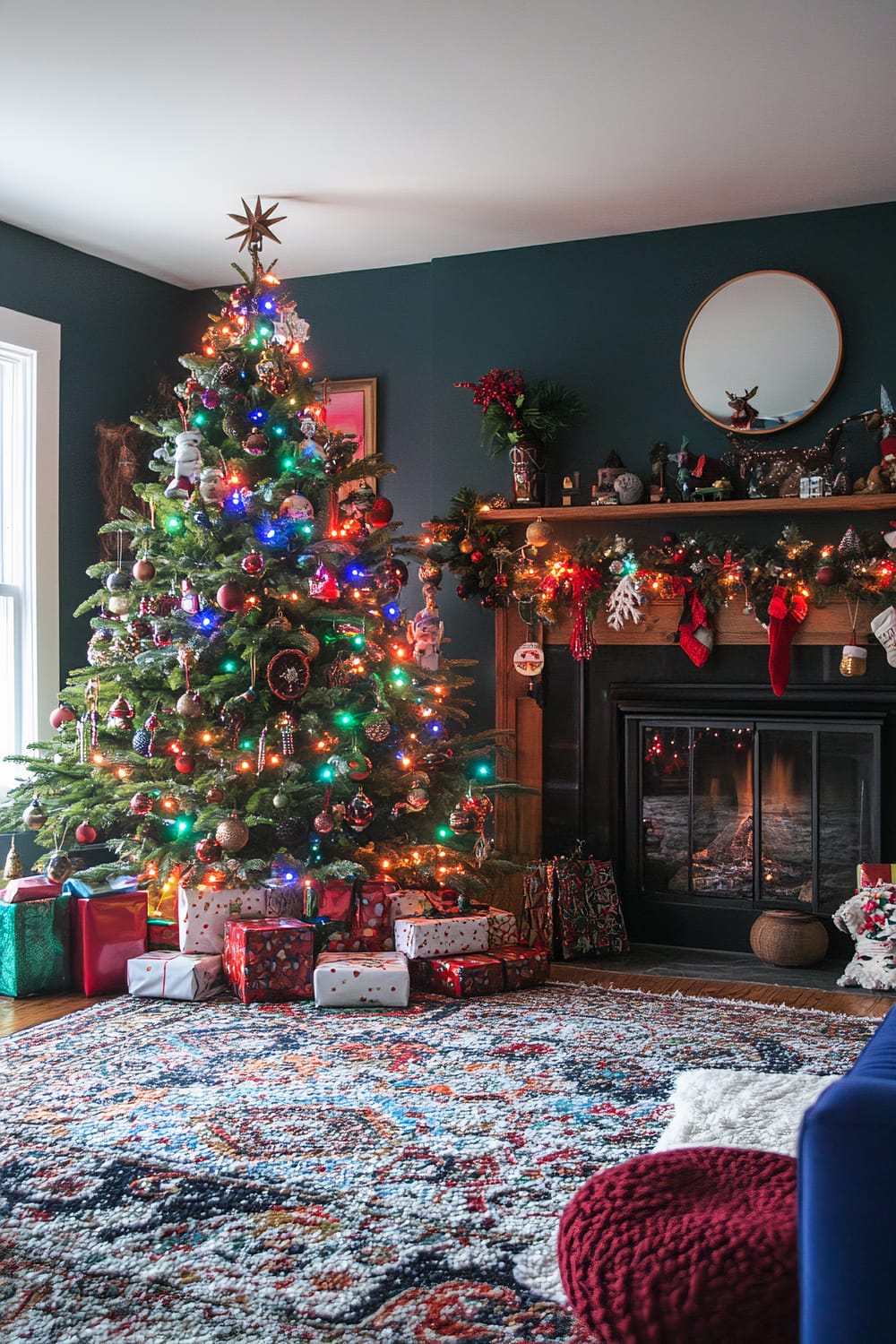 A festive living room is decorated for Christmas. A lush green Christmas tree adorned with multicolored lights, ornaments, and a star topper stands prominently in the corner. Underneath the tree is an array of wrapped gifts in various colors. To the right, a fireplace with a roaring fire is decorated with evergreen garlands, red stockings, and festive knickknacks. Above the wooden mantel is a round mirror reflecting part of the room. A cozy patterned rug covers the floor, with a plush red ottoman and part of a blue sofa visible in the foreground.