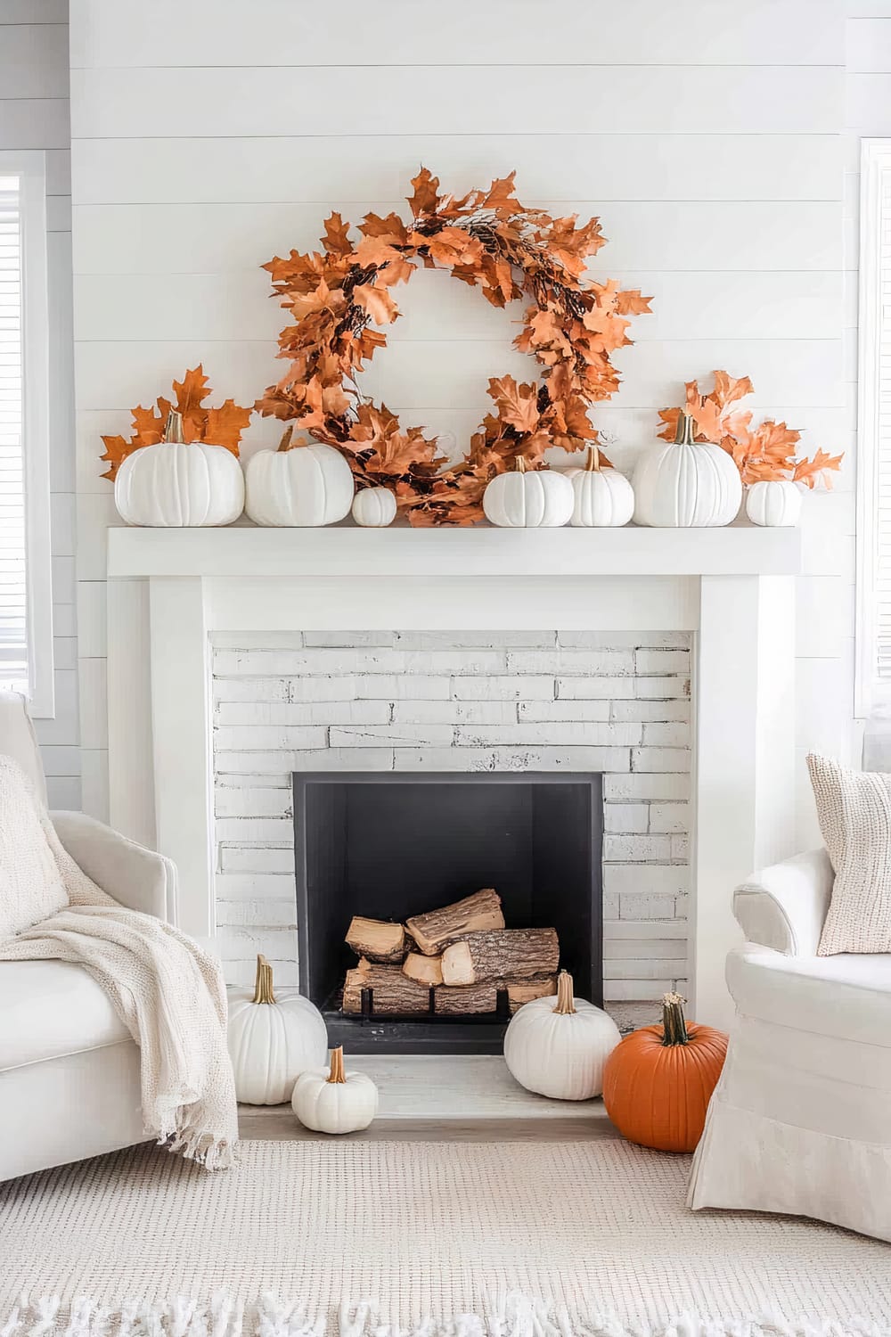 A bright, tastefully decorated fireplace mantel scene showcases a modern white aesthetic with autumnal accents. The wall features horizontal white shiplap paneling. On the mantel, a combination of white and one orange pumpkin are interspersed among an ornate wreath of orange, fall-colored maple leaves. Below the mantel, the fireplace is framed in white brick with a black interior holding a few logs, ready for a fire. White pumpkins, both large and small, surround the fireplace on the floor. Two white upholstered armchairs, each draped with a knitted throw blanket and positioned on a light-colored woven rug, frame the scene.