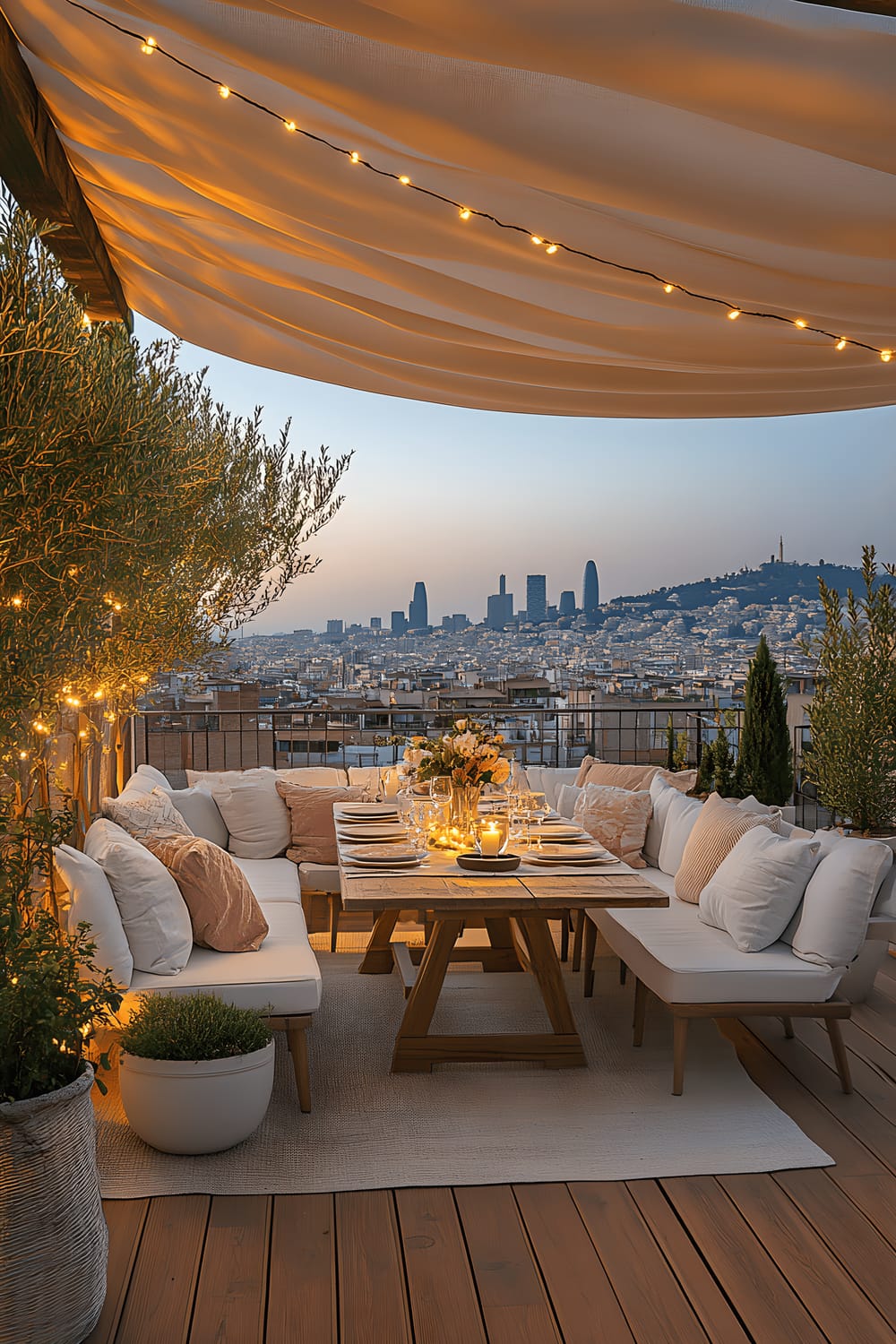 A wide-angle image of a spacious rooftop terrace in the evening lit by ambient string lights. The terrace has white and pastel-coloured modern outdoor furniture including cushioned seats and a central wooden dining table set for outdoor dining. Greenery with potted olive trees and bougainvillea plants adds a natural touch, while the background reveals panoramic views of Barcelona’s skyline, featuring landmarks like Sagrada Família and Montjuïc.