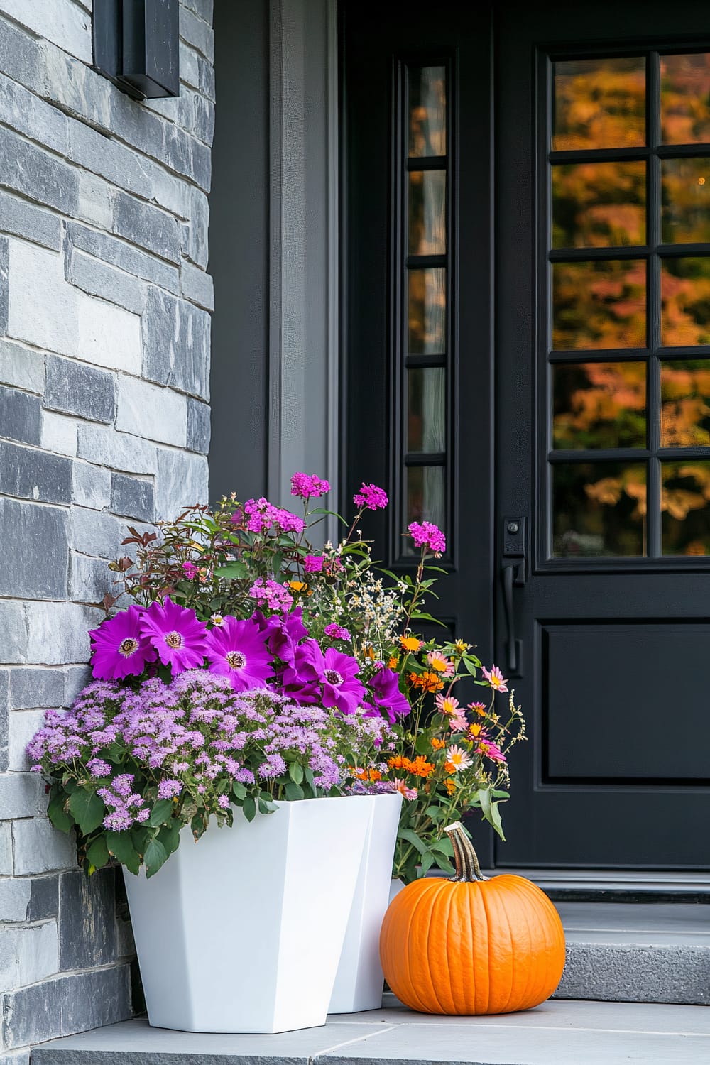 A front doorstep decorated with a vibrant flower arrangement in a white planter and an orange pumpkin. The planter contains a mix of purple, pink, and orange flowers, along with greenery. There is a modern black door with grid windows and a light grey stone wall beside the door.