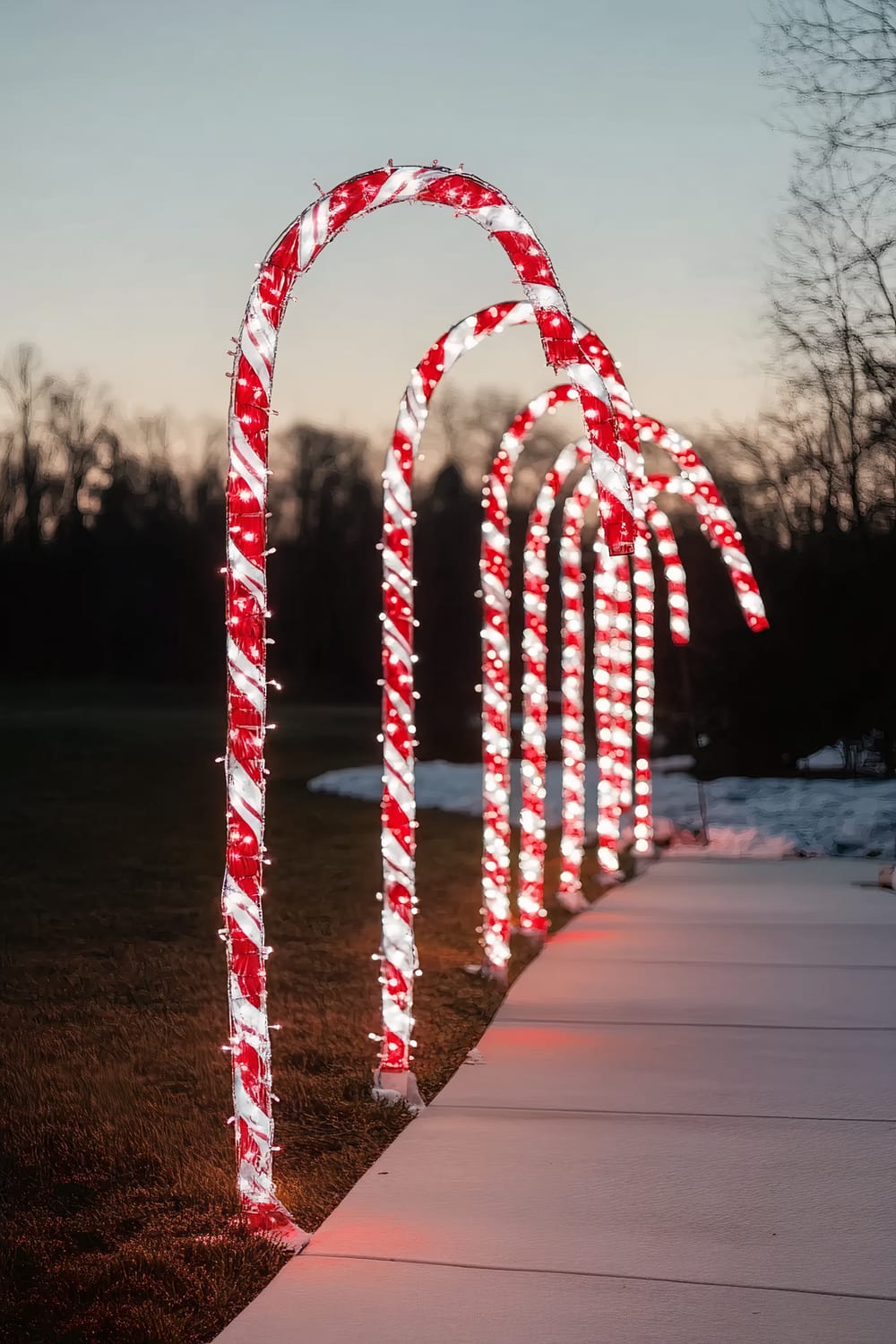 An outdoor walkway decorated with illuminated candy cane light arches. The candy canes, glowing in festive red and white lights, form a whimsical path under a twilight sky. The surrounding landscape is peaceful and shows a mix of grass and some snowy patches.