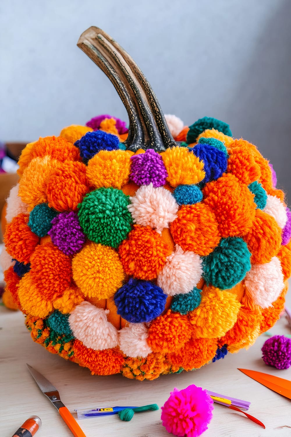 A pumpkin decorated with various colorful pom-poms. The pumpkin's stem is visible and the pom-poms are densely packed, covering the pumpkin in shades of orange, green, yellow, purple, blue, and pink. Crafting tools and materials such as scissors, yarn, and needles are scattered around it on a light wooden surface.