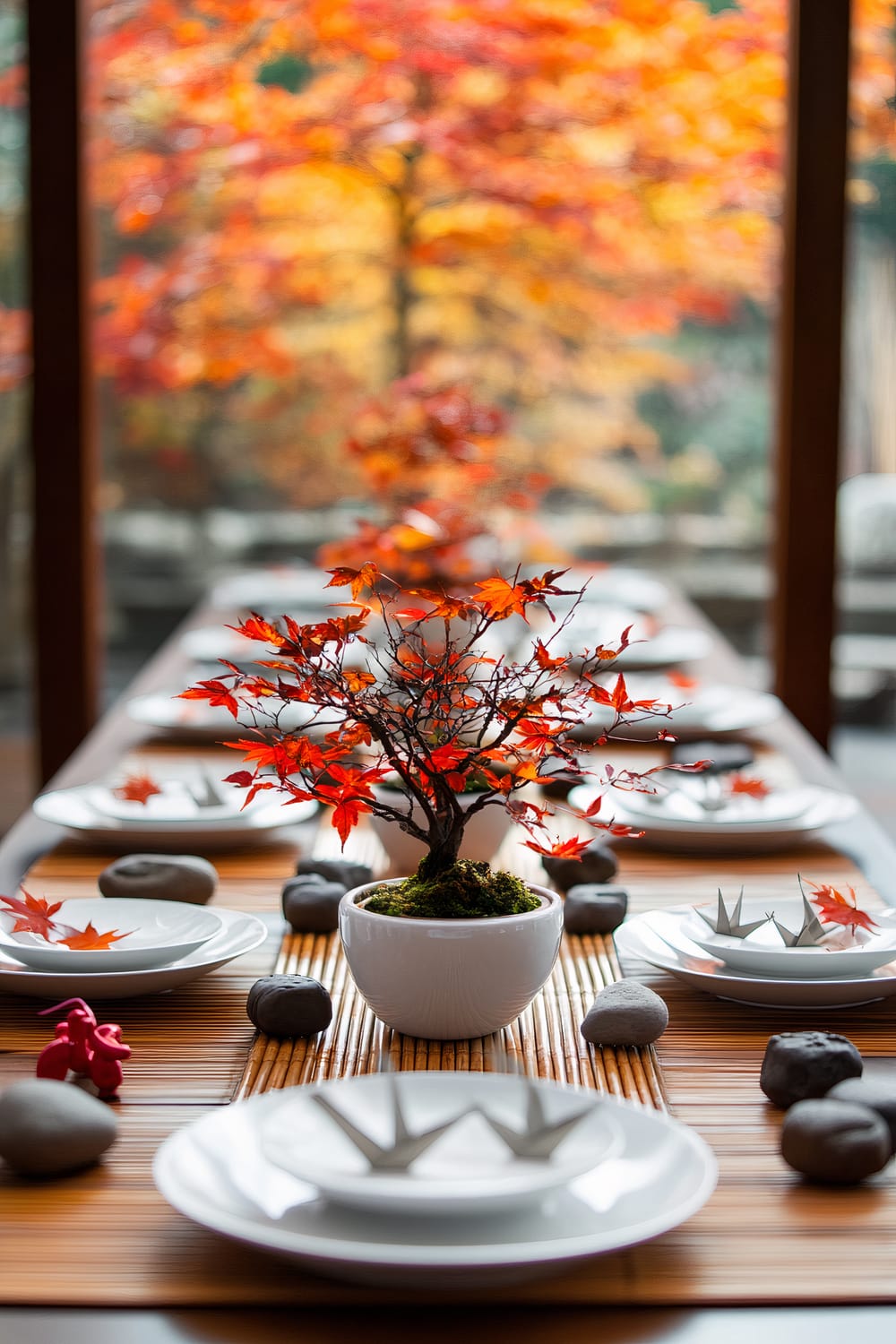 A dining table with a centerpiece of a small potted maple tree with vibrant red leaves. The table is set with white plates, gray stones, and artistic folded paper pieces. The background showcases a large glass window with a view of trees boasting autumnal colors in red, orange, and yellow.