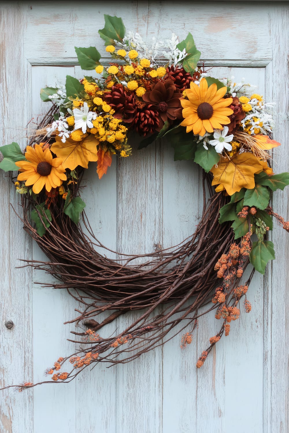 A decorative wreath composed of twisted brown branches, adorned with various artificial flowers and foliage. Prominent elements include bright yellow sunflowers, small white flowers, yellow clusters, green leaves, brown pinecones, and orange berries. The wreath is mounted on a weathered, light blue wooden door, providing a rustic and autumnal aesthetic.