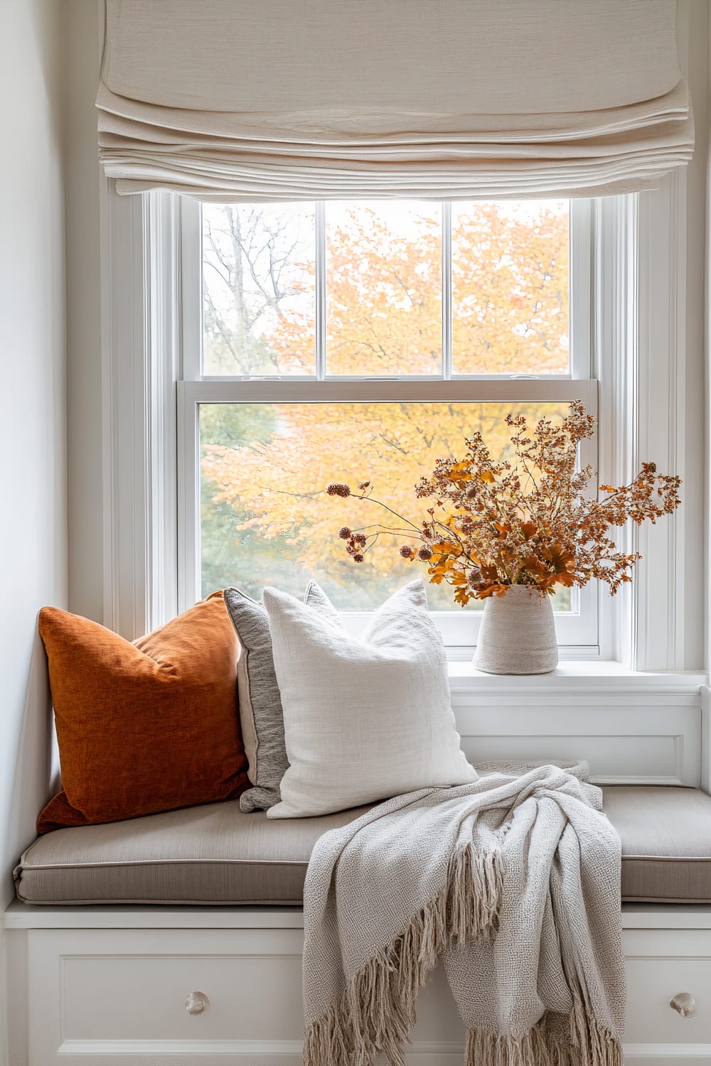 A serene window nook featuring a cushioned seating area with a beige mattress, complemented by a burnt orange pillow and two white pillows. A soft gray throw with fringed edges is draped over the side. The window overlooks a scene with trees showcasing autumn foliage, creating a beautiful backdrop. A vase filled with orange and brown dried flowers sits on the windowsill, adding a touch of fall decor. Cream-colored Roman shades hang above the window, providing a warm, calming atmosphere.