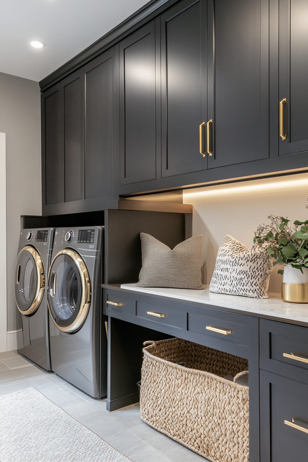 A modern laundry room featuring sleek black cabinetry with gold handles, an under-counter washer and dryer with gold accents, a countertop with two decorative pillows, and a woven storage basket underneath.