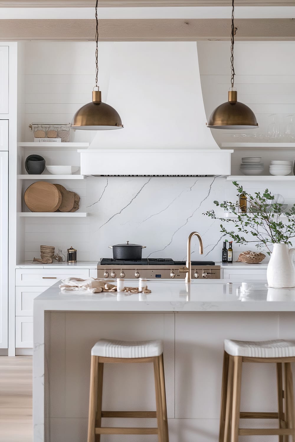 A modern kitchen featuring a central island with a white marble countertop and two wooden stools with white cushioned seats. Above the island, two bronze pendant lights hang from the ceiling. In the background, there's a white stovetop with an oven, and a sleek white range hood above it. The backsplash is made of white marble with subtle grey veining. Open shelves flanking the range hood hold various dishes and decorative items, including wooden cutting boards and glass bottles. A gold faucet is installed on the island.