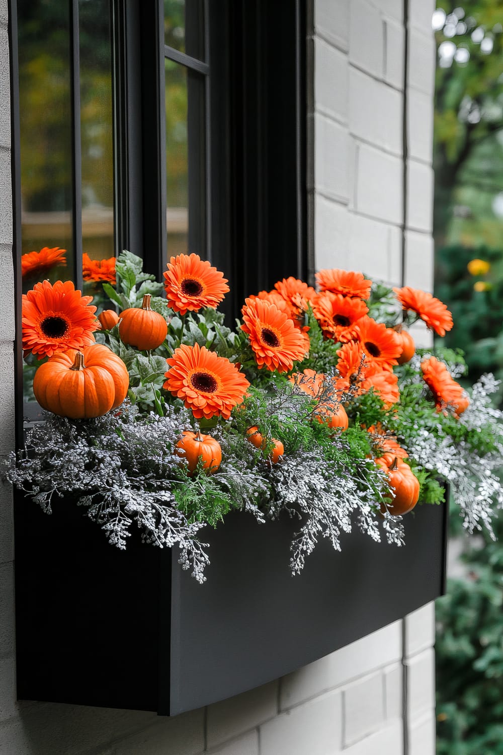 A close-up of a window box attached to the exterior wall of a house. The black window box is filled with vivid orange gerbera daisies, small orange pumpkins, lush green foliage, and branches covered in white frost. The window above has a black frame and is partially opened, reflecting the orange flowers and green surroundings. The backdrop is a light-colored brick wall and blurred greenery from the adjacent garden.