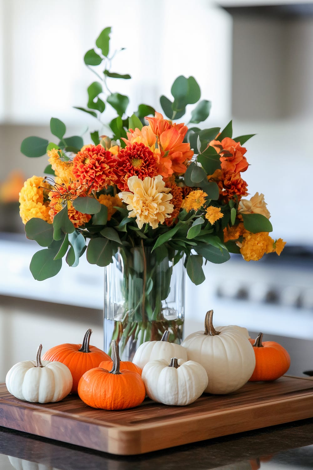 A close-up of a wooden cutting board with multiple small pumpkins in white and orange hues. Behind the pumpkins is a tall, clear glass vase filled with an assortment of vibrant orange, yellow, and cream-colored flowers, along with green foliage.